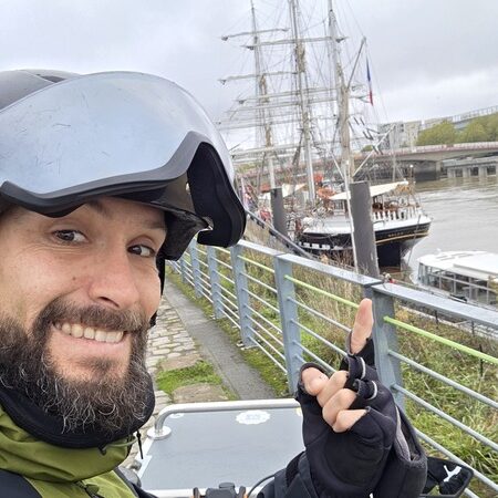 Thomas Nataf électricien à vélo centre ville de Nantes devant le Belém à quai, il porte son casque avec visere et souri à l'objectif. Derrière le pont anne de bretagne en travaux en 2025