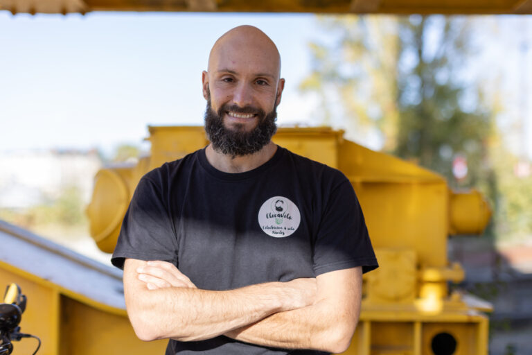 Elecavelo assis sur la caisse de son vélo sous la grue jaune les bras croisés, souriant